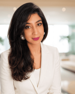 Meet our Experts: A woman with long dark hair and arched eyebrows faces the camera indoors, with blurred lights and windows in the background.